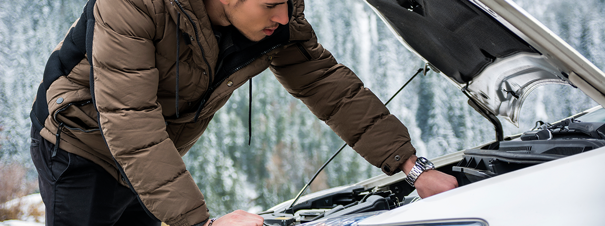 Man looking under the hood of his car on the side of the road during winter